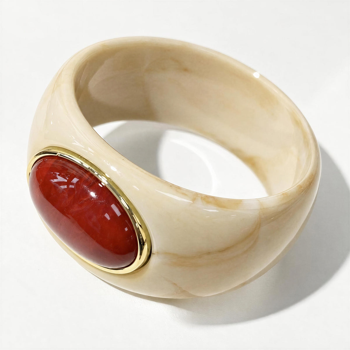 The Bruna Bangle from Chica Chica Jewelry.  A wide, chunky bangle ring made of cream-colored marbled resin, featuring a large, polished oval red stone in a gold bezel setting on the front, photographed on a white background.
