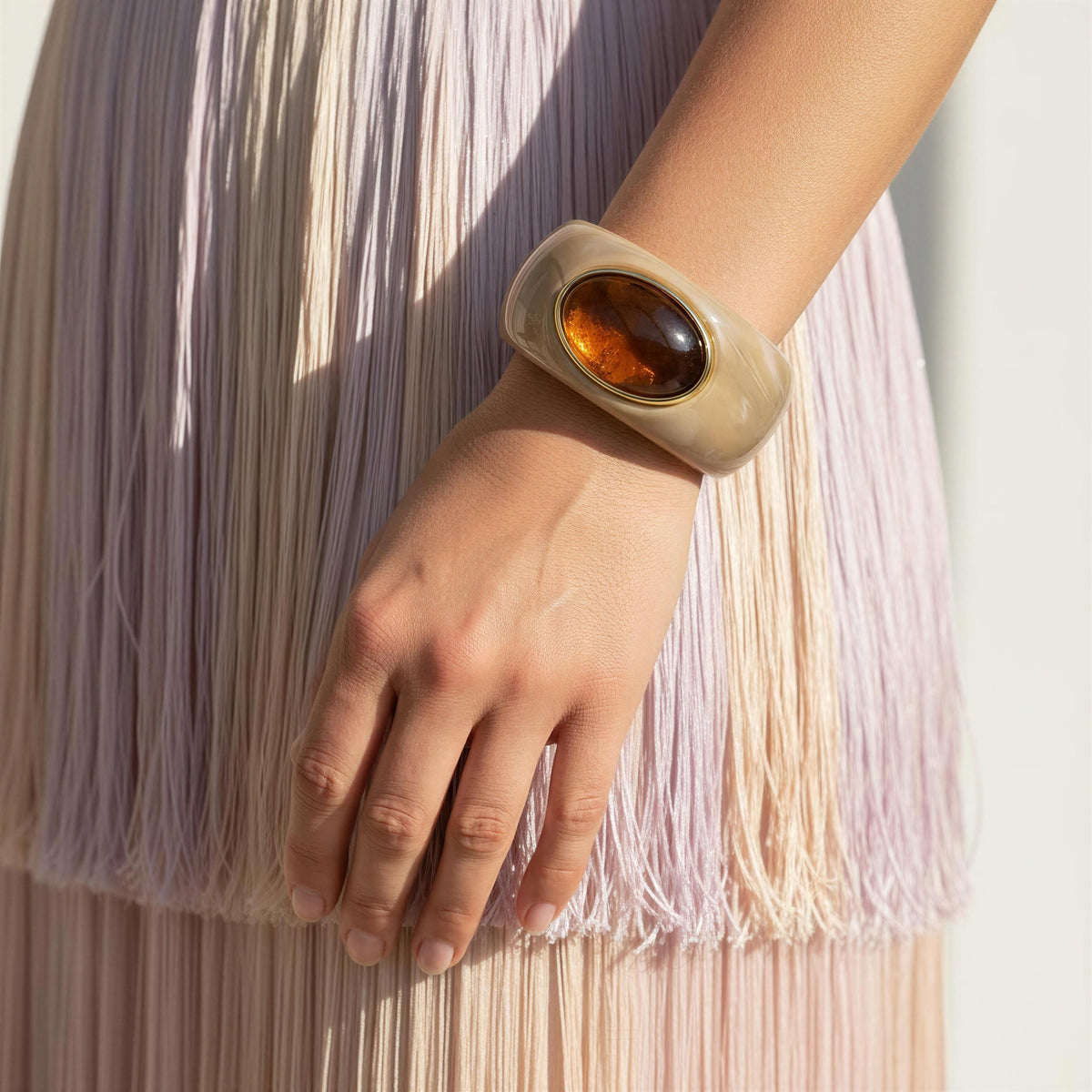 A close-up of a person's hand and wrist wearing the  Bruna Bangle from Chica Chica Jewelry,  a chunky cream-colored cuff bracelet with a polished amber oval centerpiece, set against a soft pastel fringed dress.