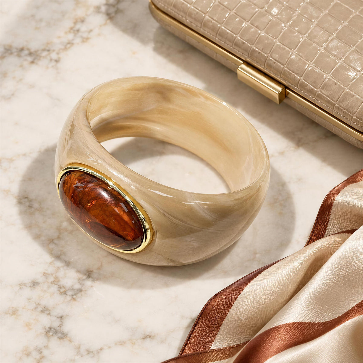 The Bruna Bangle from Chica Chica Jewelry.  A chunky bangle bracelet made of marbled cream-colored resin with a polished amber-brown oval inset and gold-tone bezel, styled on a marble surface next to a clutch and satin ribbon.