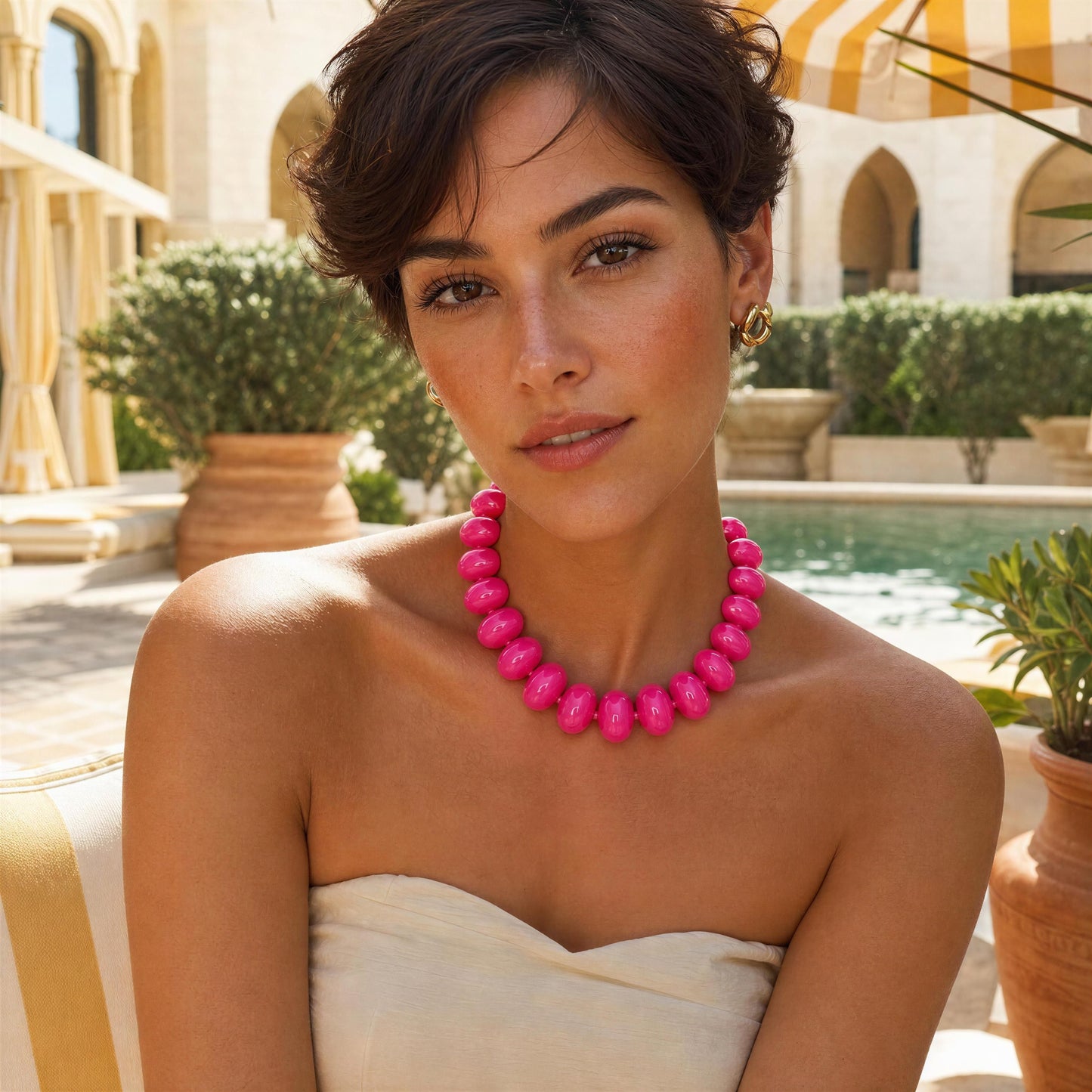 Woman wearing a bright pink necklace in an outdoor setting with a pool and building in the background.