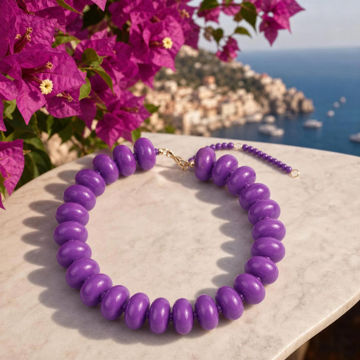 The Capri Lilla Choker, a purple beaded necklace with large oval beads and a gold clasp, displayed on a light marble surface with a scenic coastal background and bright pink flowers.