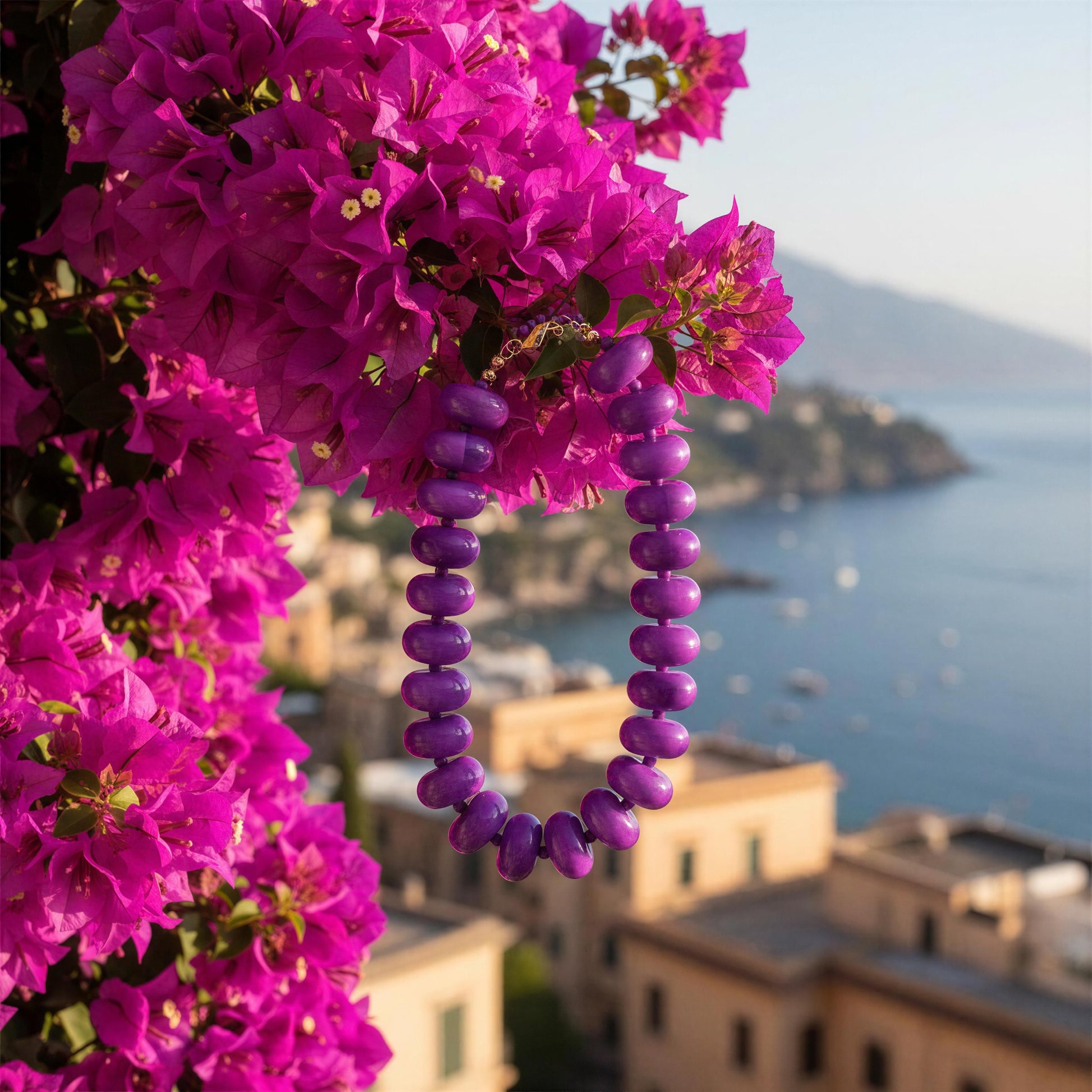 The Capri Lilla Choker draped over bright magenta bougainvillea flowers, with a blurred coastal town and sea in the background.