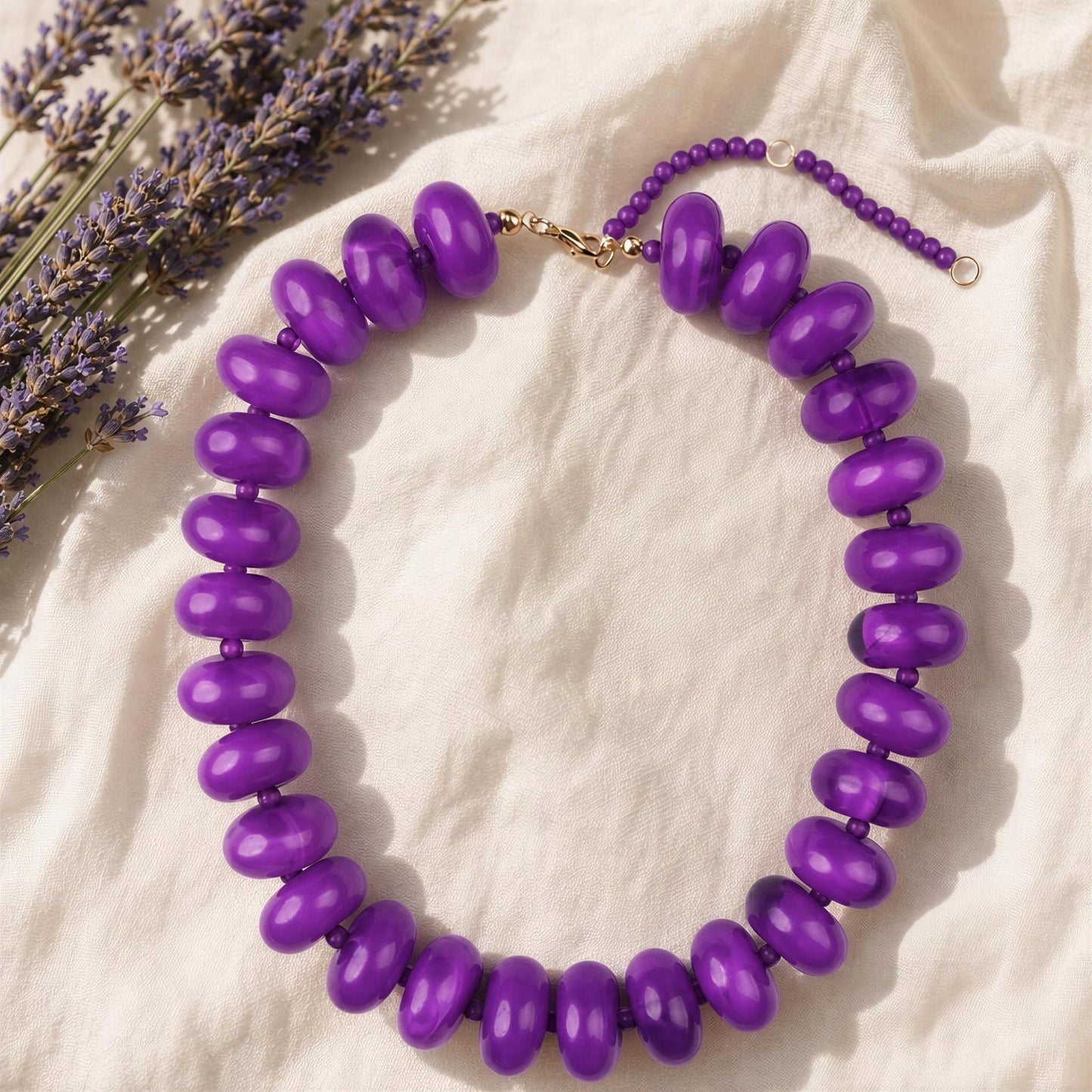 A purple beaded necklace with large, glossy, rounded beads and a gold-tone clasp, displayed on a textured cream fabric next to dried lavender sprigs.