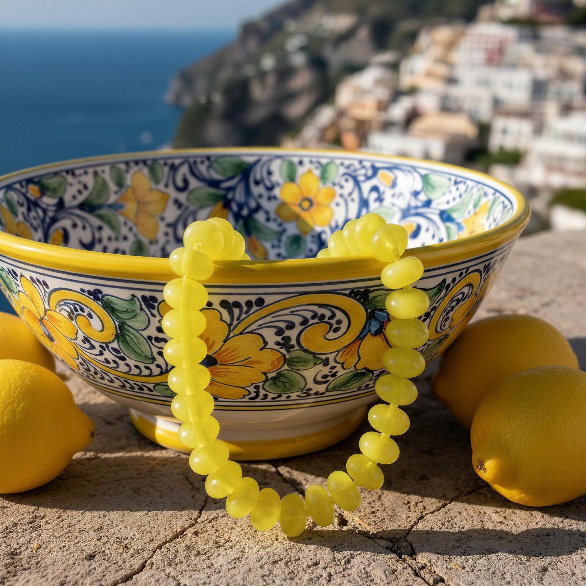 The Capri_Limone_Choker from Chica Chica Jewelry draped over a colorful ceramic bowl, with lemons on either side and a blurred coastal town and sea in the background.