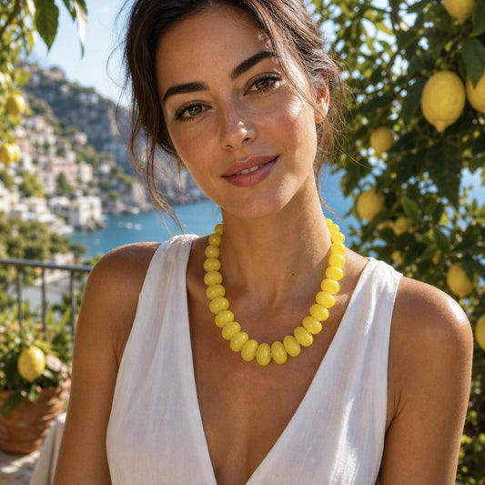 A woman wearing a chunky yellow beaded necklace from Chica Chica Jewelry with a white sleeveless top, set against a sunny outdoor background of lemon trees and a coastal sea view.