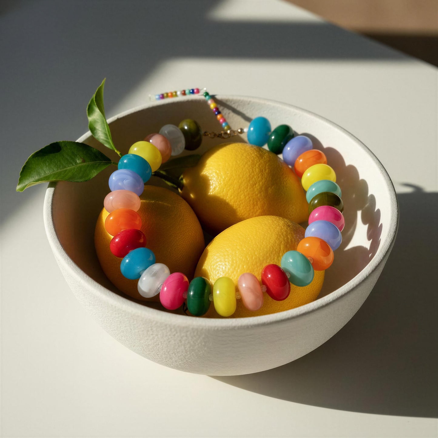 A white ceramic bowl filled with bright yellow lemons and a colorful beaded bracelet, photographed on a sunlit white surface with strong shadows.