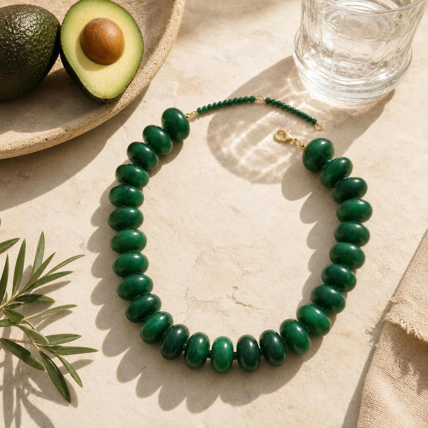 Green beaded necklace on a light surface with an avocado and glass of water in the background