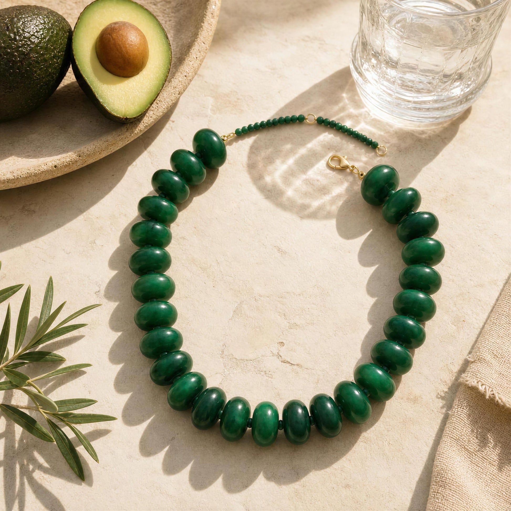 Green beaded necklace on a light surface with an avocado and glass of water in the background