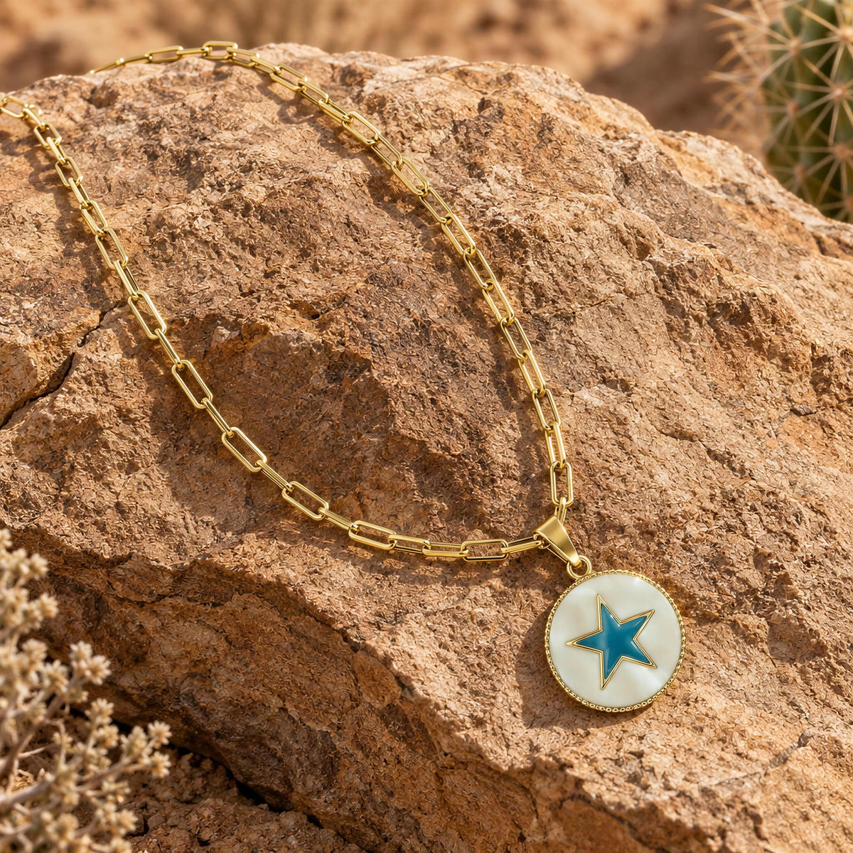 The  Celestia Turquoise Star Necklace- Chica Chica Jewelry, gold paperclip chain necklace with a round pendant featuring a blue star on a white background, displayed on a large brown rock outdoors.