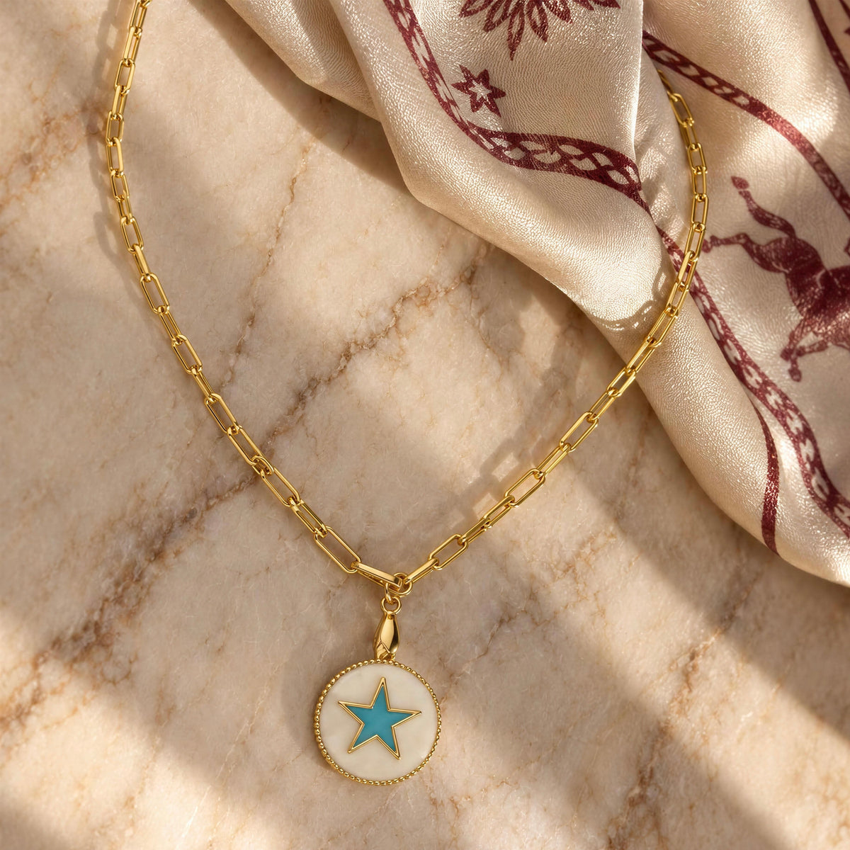 A gold paperclip chain necklace with a round pendant featuring a white enamel background and a light blue star, styled on a beige marble surface next to a cream silk scarf with a burgundy deer pattern.