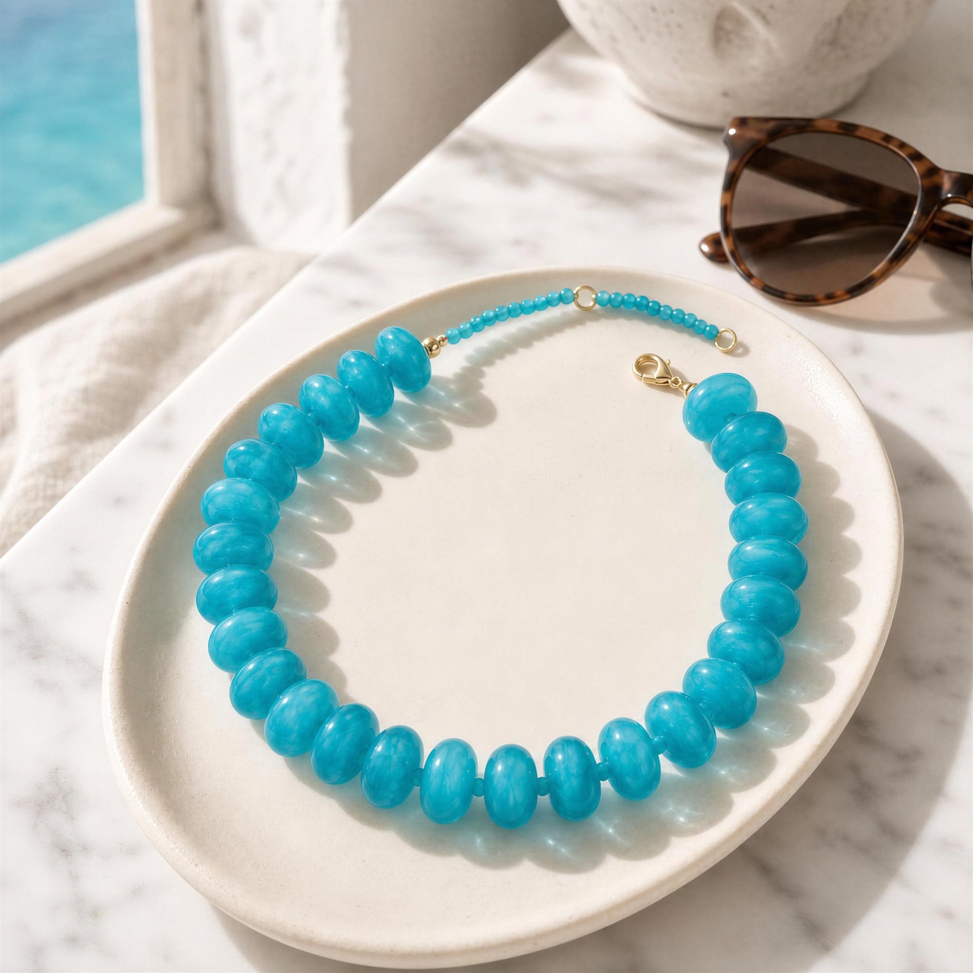 Turquoise beaded necklace on a white plate with sunglasses and a marble surface.