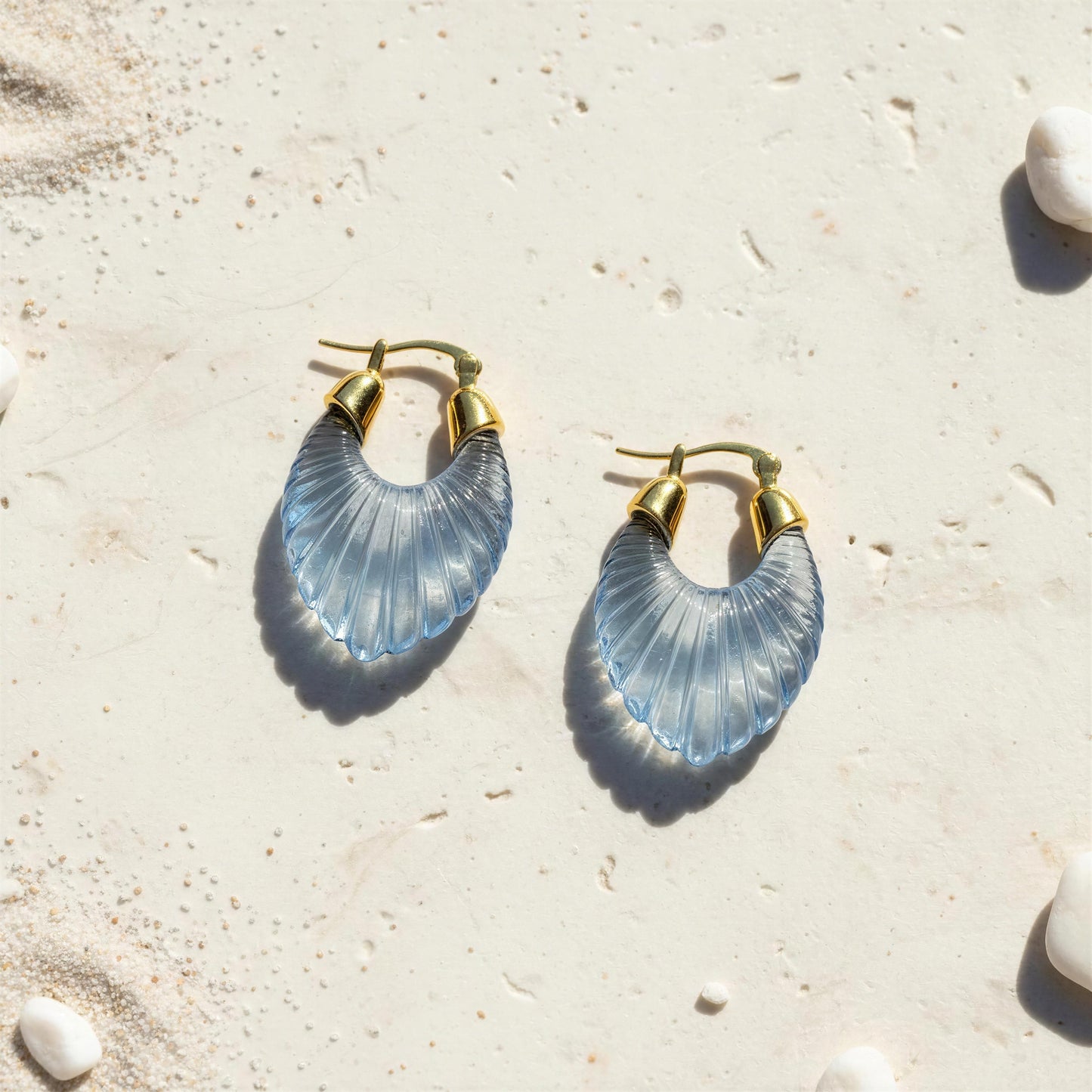 A pair of gold-tone hoop earrings with translucent light blue shell-shaped dangles, displayed on a sandy surface with small white shells.