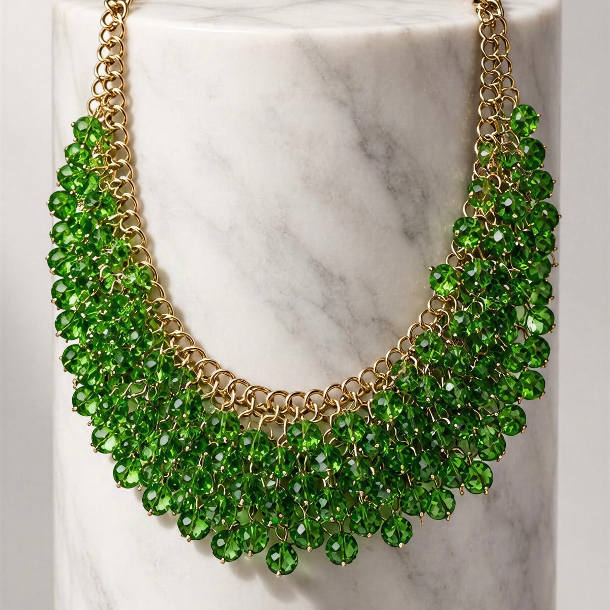 The Eleonora Necklace from Chica Chica Jewelry.  A gold-tone statement necklace with multiple rows of faceted green gemstones, displayed on a white marble cylinder.