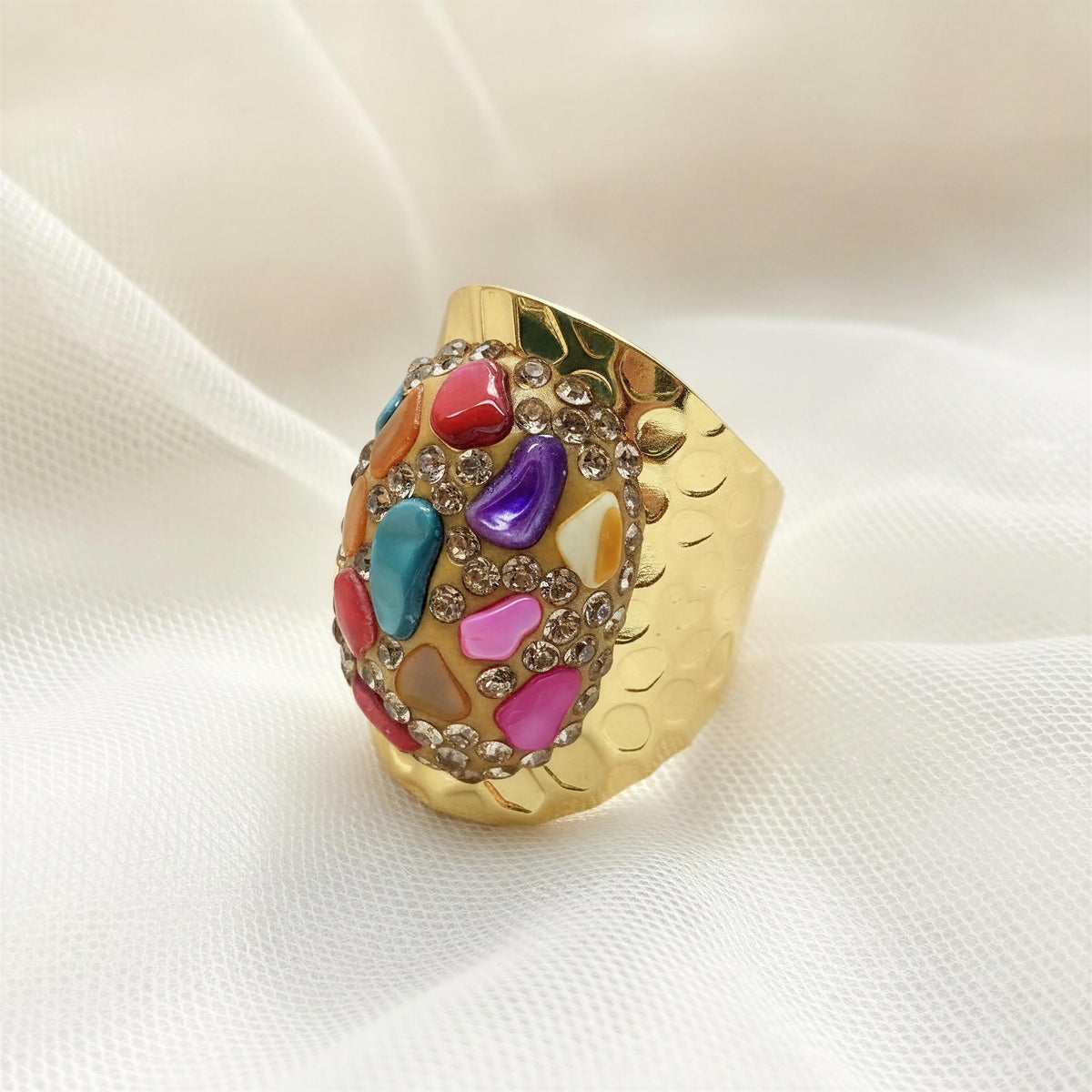 The Chroma Ring from Chica Chica Jewelry.  A wide gold ring featuring a colorful mosaic of faux gemstones in pink, blue, purple, red, and orange, accented with small clear rhinestones, displayed on a soft white fabric background.