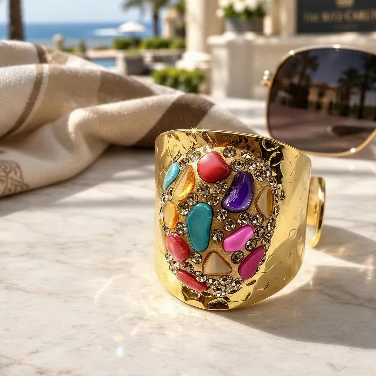 The Chroma Ring from Chica Chica Jewelry, a  wide gold ring with a hammered finish, decorated with a colorful mosaic of faux gemstones in pink, blue, purple, yellow, and red, set on a marble surface with sunglasses and a beach background.
