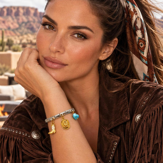 A woman in a brown suede fringe jacket wearing a silver-toned charm bracelet with gold and turquoise details, posed against a desert canyon background.