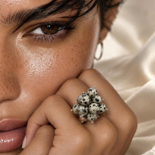 A close-up lifestyle shot of a model wearing the Dalma Ring from  Chica Chica Jewelry, a  large silver ring adorned with a cluster of white and black spotted beads, resting her chin on her hand against a soft cream satin background.