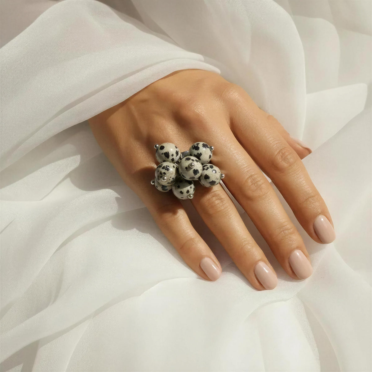 A close-up of a hand wearing the Dalma Ring from Chica Chica Jewelry -  a cluster ring made of white beads with black spots, styled with a flowing white fabric background.