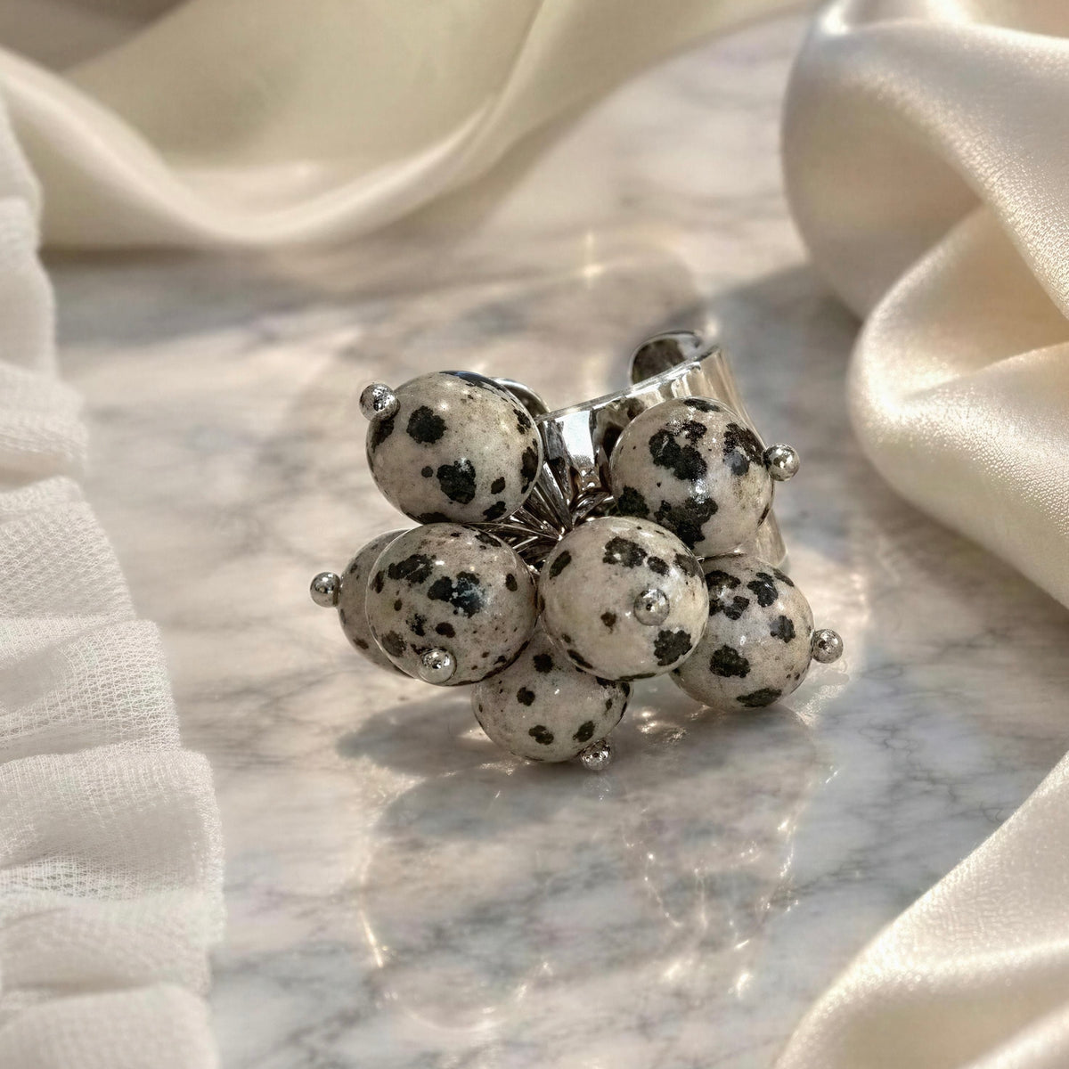 The Dalma Ring  from Chica Chica Jewelry, a  silver ring featuring a cluster of black-and-white spotted stone beads, styled on a white marble surface with draped cream satin fabric.