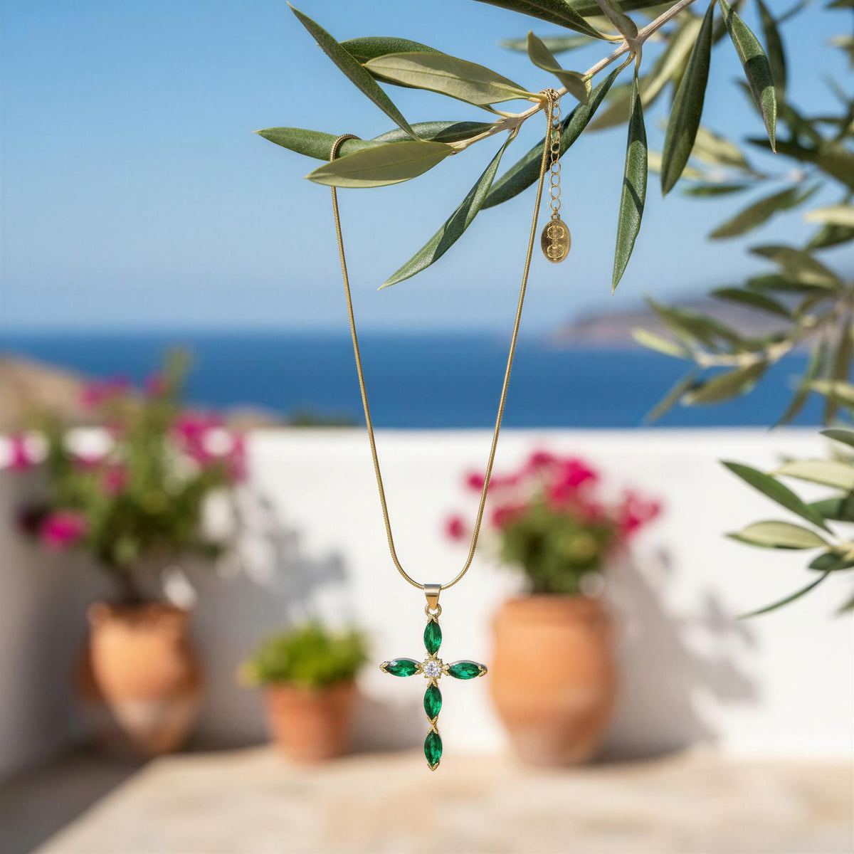 The Defne Cross Necklace from Chica Chica Jewelry; a  gold cross pendant necklace with green emerald-cut stones and a central clear gemstone, hanging from olive branches with a blurred Mediterranean background of blue sea and potted flowers.