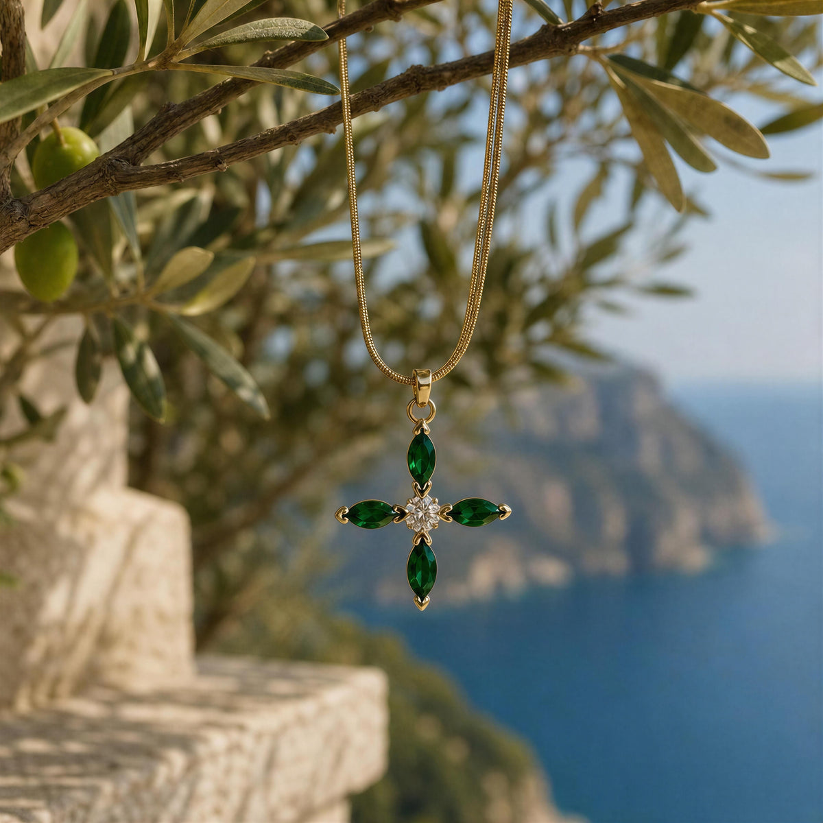 A gold cross pendant necklace with four green marquise-cut stones and a clear central stone, hanging from an olive tree branch with a scenic blue sea and cliffs in the background.