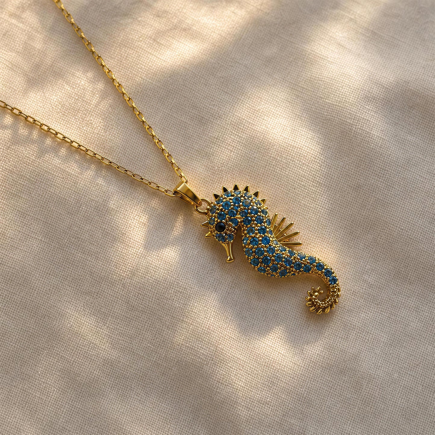 A gold seahorse pendant necklace adorned with blue and gold pavé stones, resting on a textured beige linen fabric with soft leaf shadows.