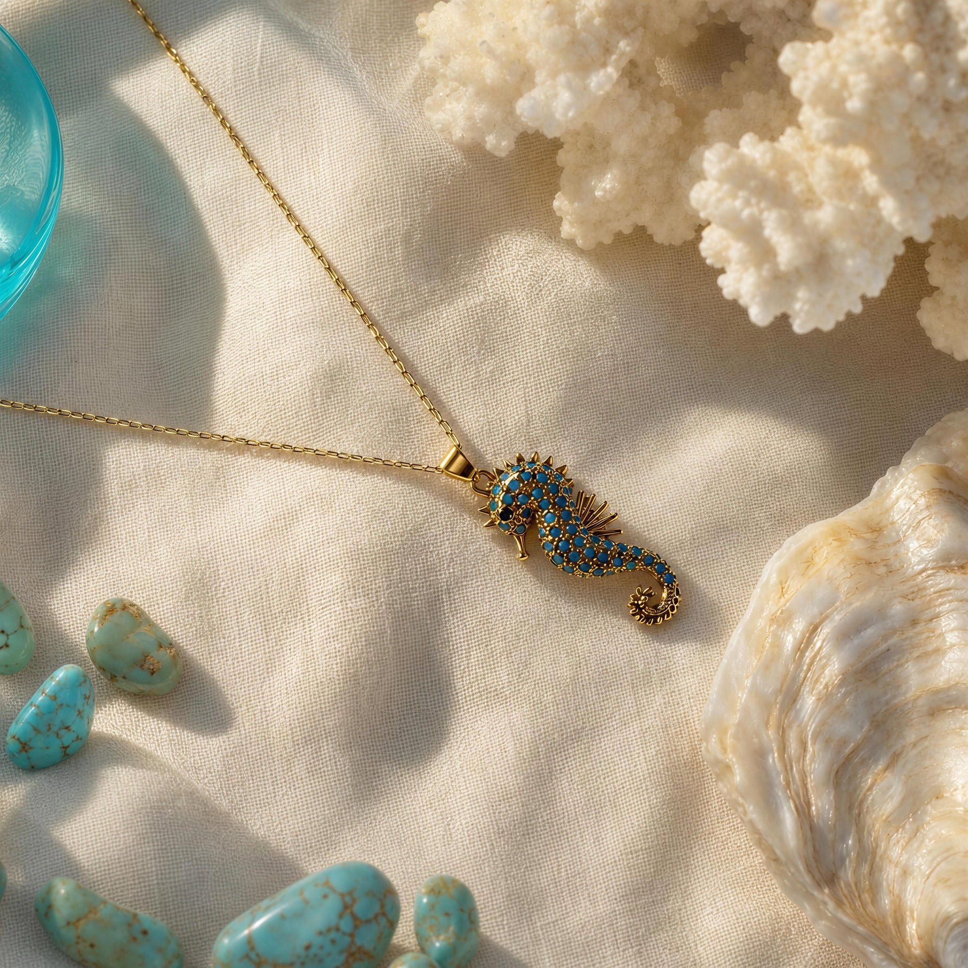 A gold necklace with a seahorse pendant embellished with blue stones, styled on a textured beige fabric with seashells, turquoise stones, and coral.