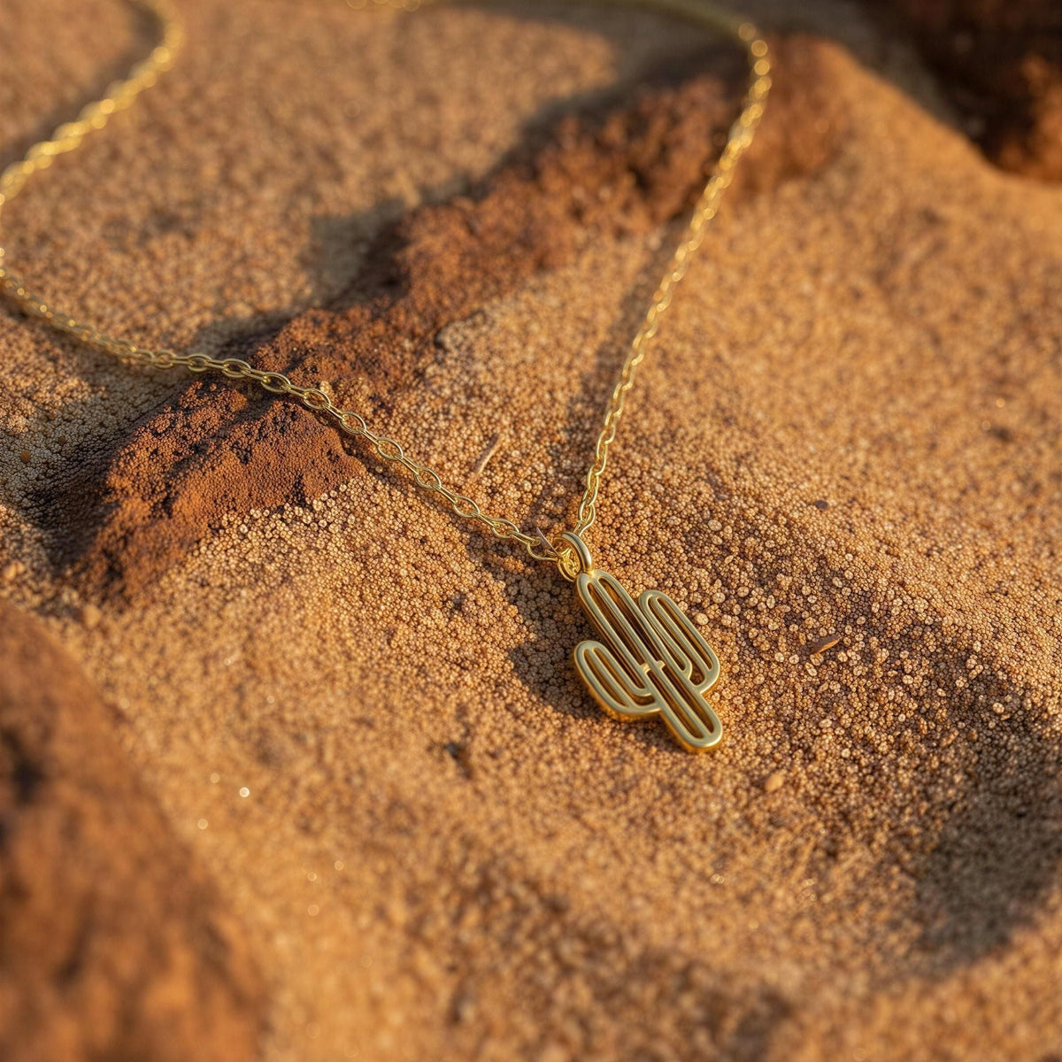 The Desert Bloom Cactus Necklace from Chica Chica Jewelry  gold cactus pendant necklace resting on textured sand with warm golden-hour lighting.