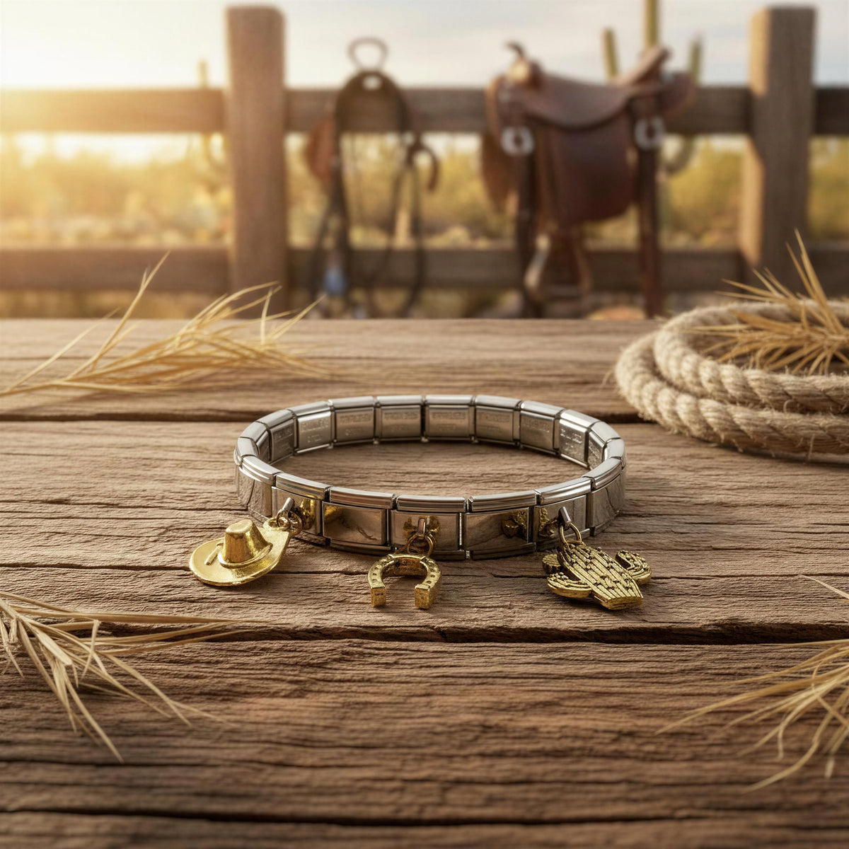 A silver-toned elastic charm bracelet with gold-toned western-themed charms (a cowboy hat, horseshoe, and hamsa hand) resting on a rustic wooden table with straw and a rope, with a blurred fence and saddle in the background.