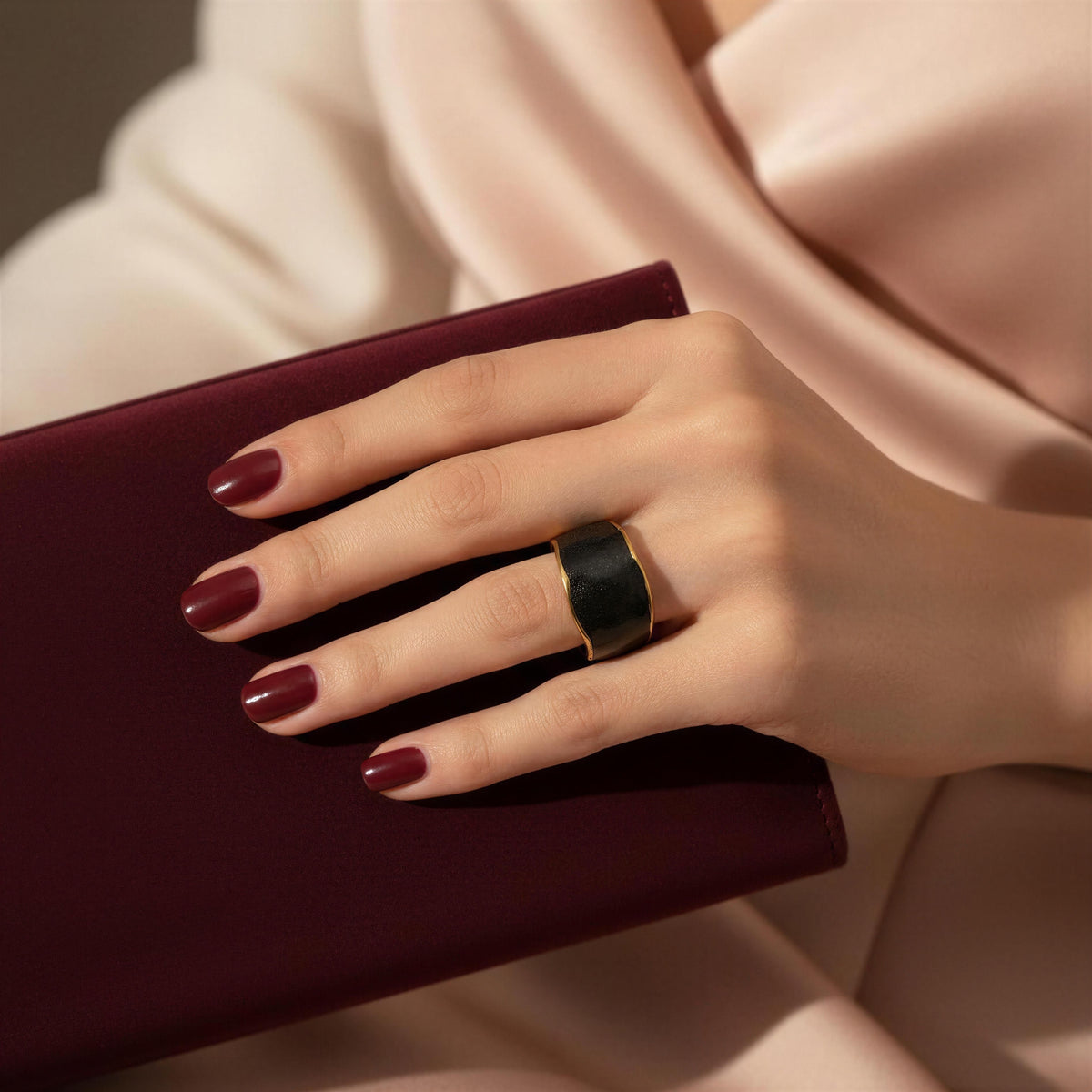 A close-up of a hand wearing the Duna Ring – Ónice from Chica Chica Jewelry, a wide black ring with gold trim, resting on a deep burgundy clutch against a soft blush pink satin background.