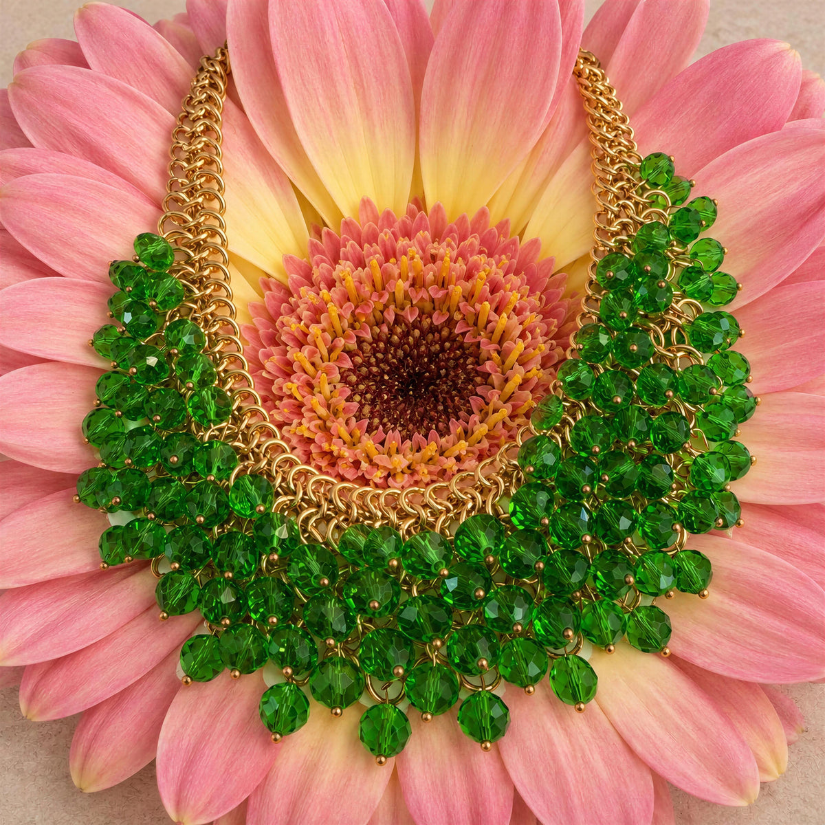 The Eleonora Necklace from Chica Chica Jewelry- 
a wide collar necklace made of green faceted gemstones and gold-tone chain, displayed on the petals of a pink and yellow gerbera daisy flower.