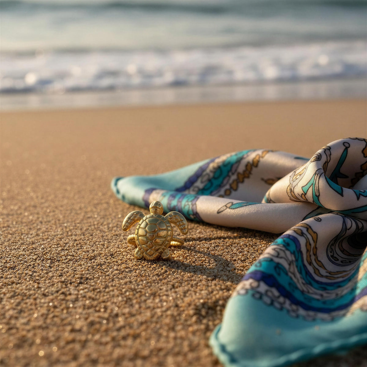 The Eternal Tortoise Ring, a small gold turtle-shaped ring sits on wet sand next to a light blue and white patterned scarf, with the ocean waves blurred in the background.