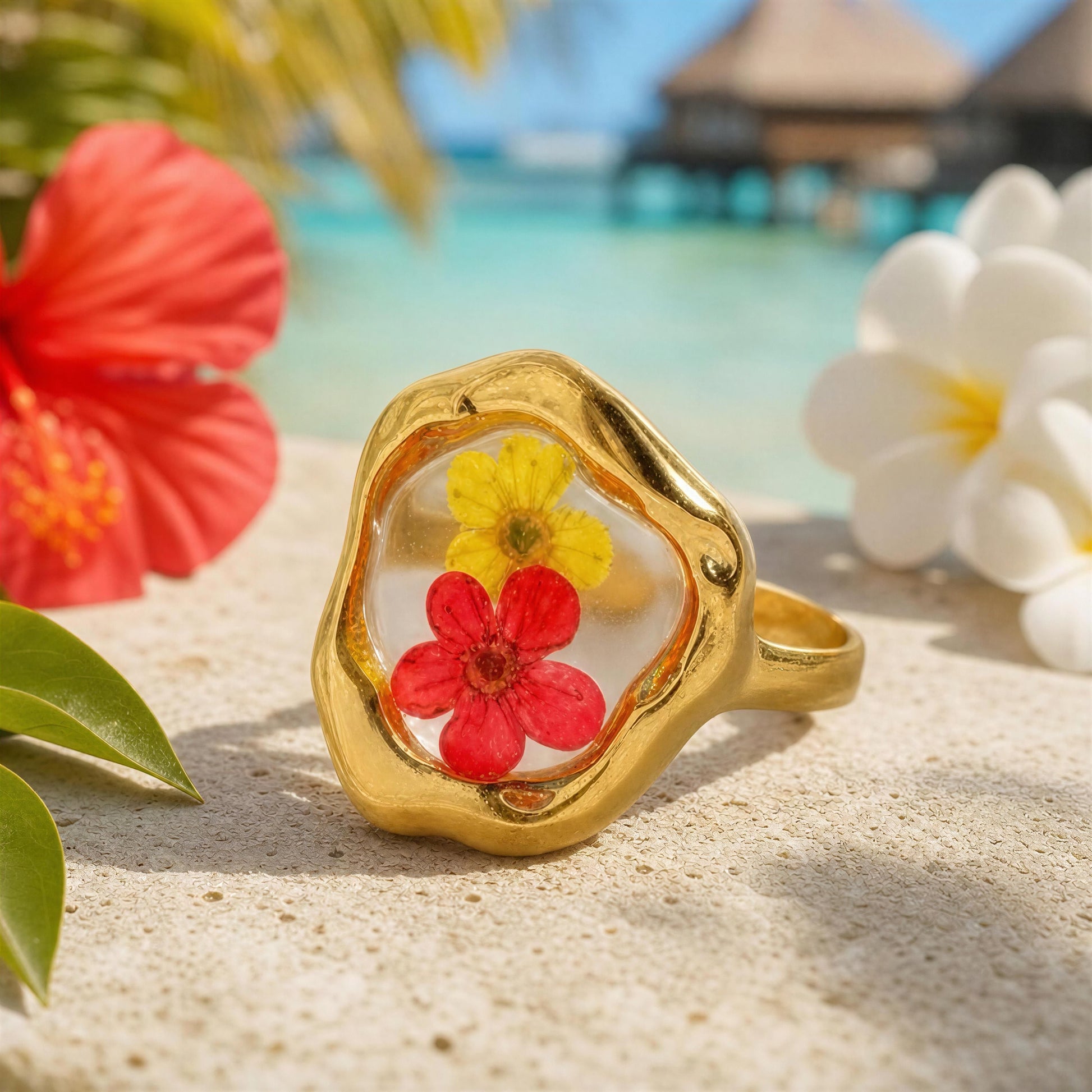 Gold ring with embedded red and yellow flowers on a sandy background with tropical flowers.