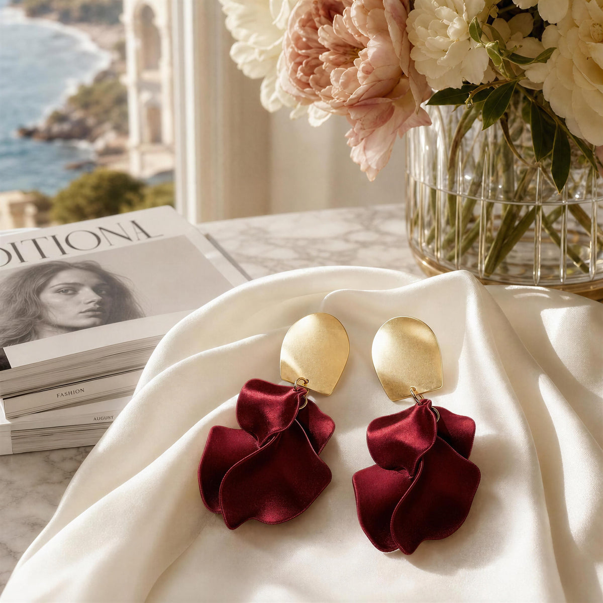 The Fiore Roja Earrings from Chica Chica Earrings- A pair of statement earrings displayed on draped white fabric, with a vase of flowers and a magazine visible in a sunlit interior setting.