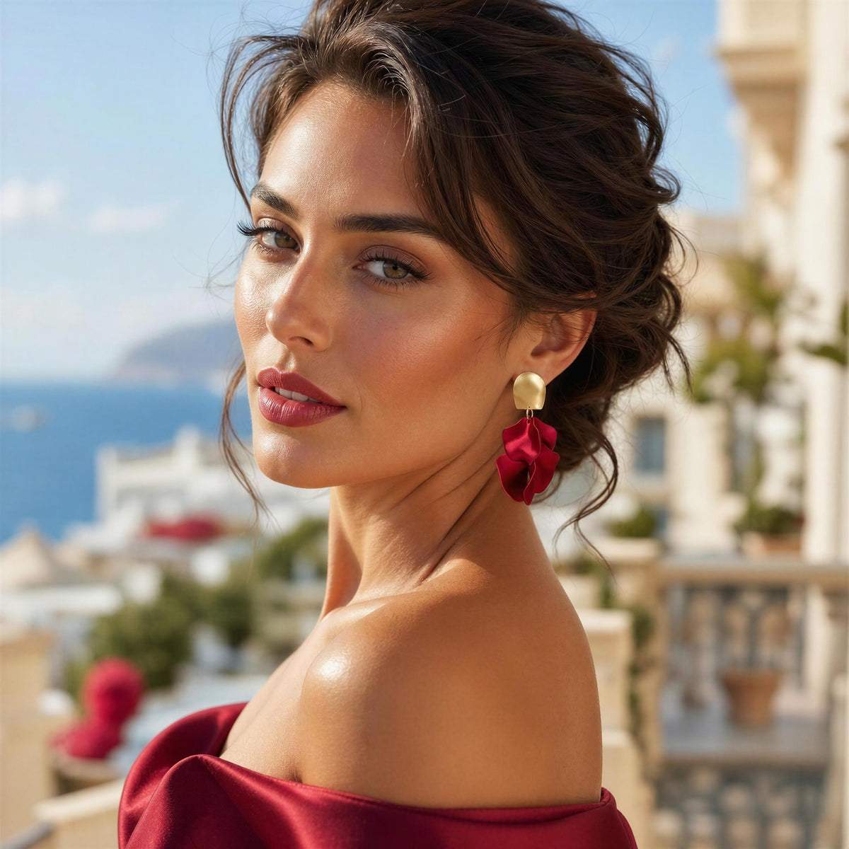 A woman with dark hair in an updo wearing a deep red off-the-shoulder top and the Fiore Roja earrings, matching red flower-shaped drop earrings with gold tops, photographed outdoors with a blurred coastal background.