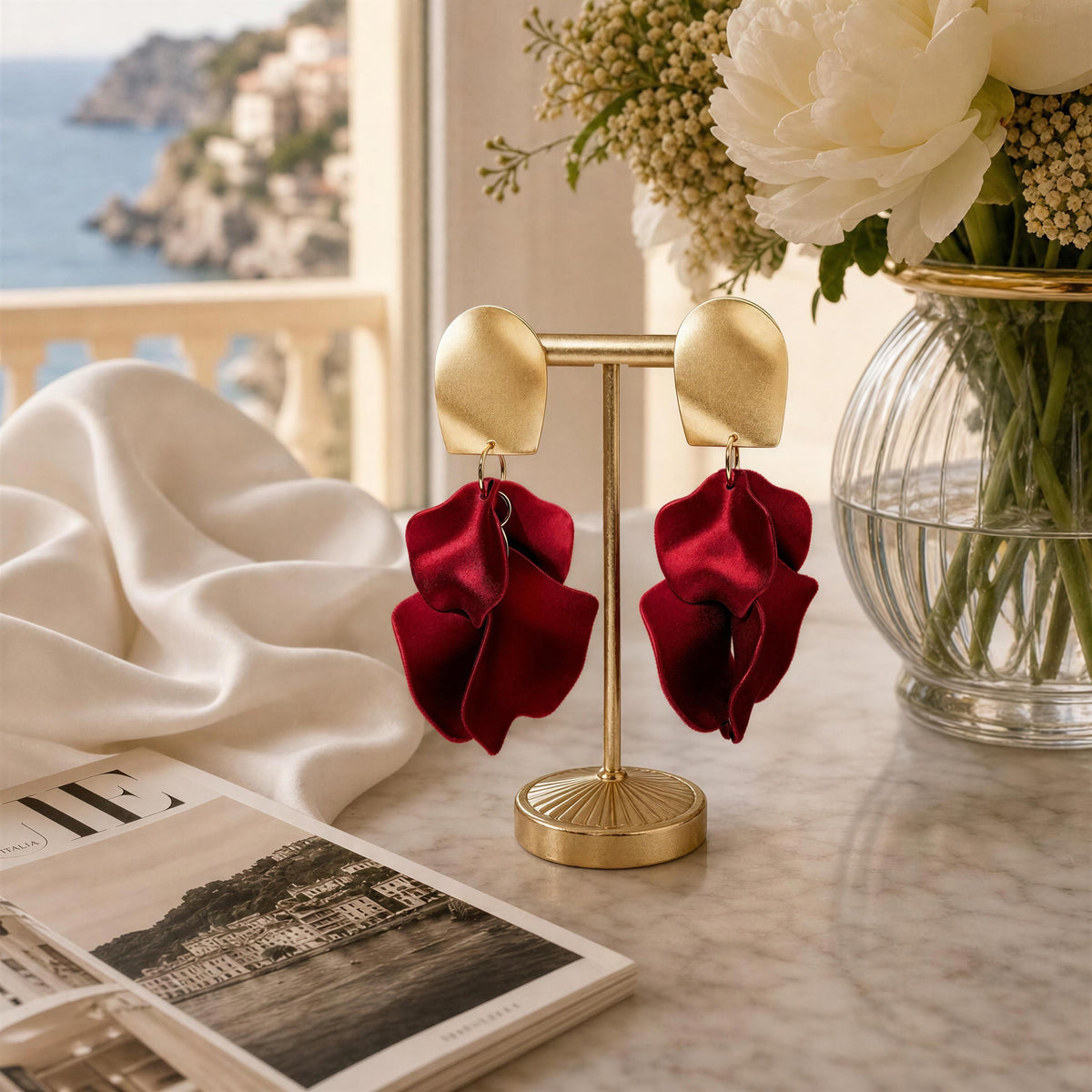 The Fiore Roja Earrings from Chica Chica Earrings. A pair of statement dangle earrings displayed on a gold stand on a marble tabletop. The earrings have matte gold oval studs and large, layered deep red satin ruffle drops.