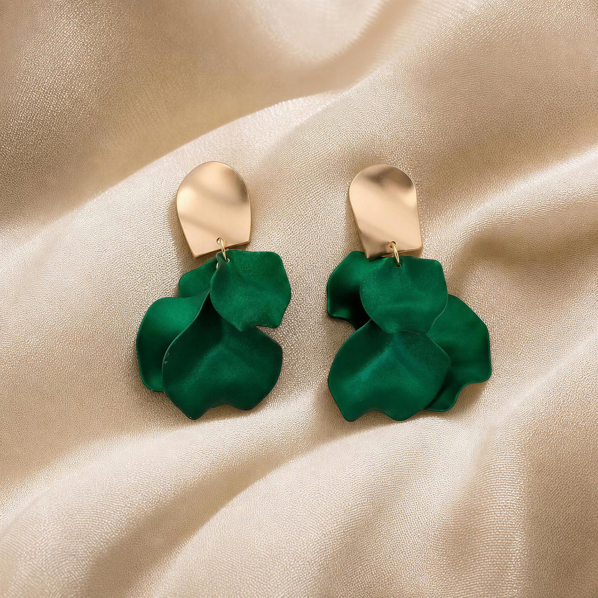 The Fiore Verde Earringa from Chica Chica Jewelry.   A pair of statement drop earrings with metallic gold-tone top pieces and layered green petal-shaped charms, displayed on draped champagne satin fabric.