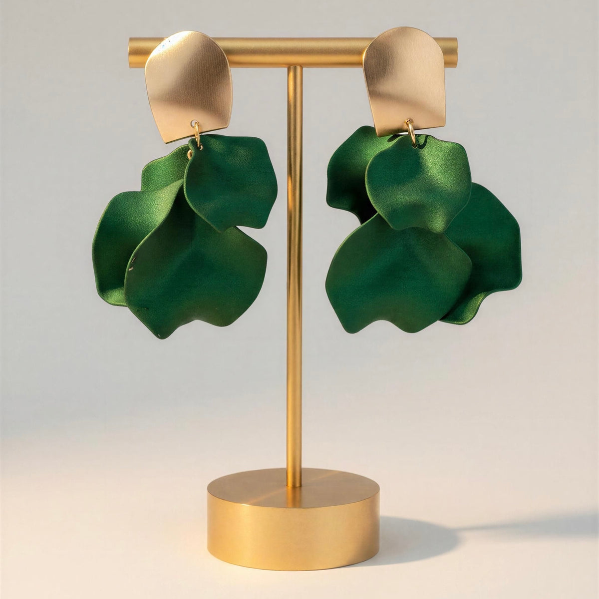 A pair of large green layered leaf-shaped dangle earrings with gold-tone top pieces, displayed on a gold stand against a light gray background.