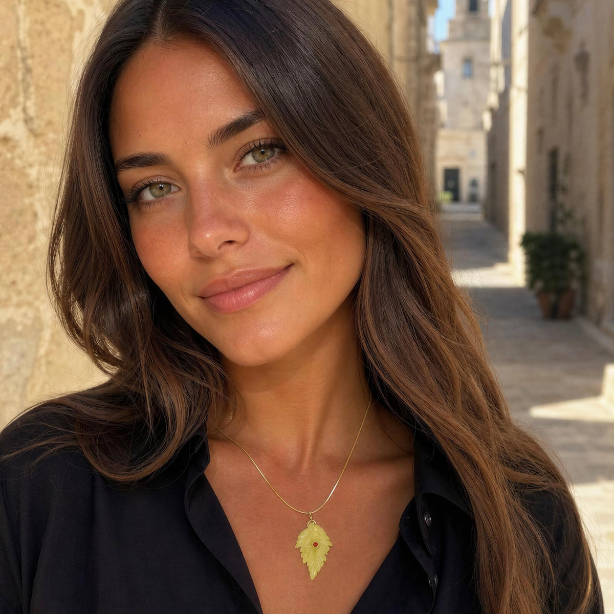 A woman with long brown hair wearing a delicate gold necklace with a leaf-shaped pendant, photographed outdoors in a sunny stone alleyway.