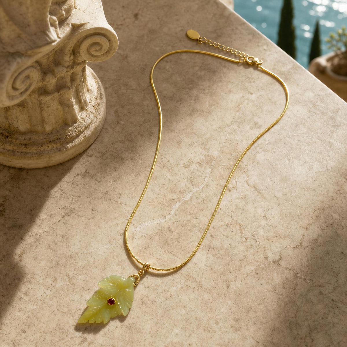 The Fiorella Necklace - Terra Collection from Chica Chica Jewelry  gold chain necklace with a carved light green leaf pendant featuring a small red gemstone, displayed on a beige stone surface.