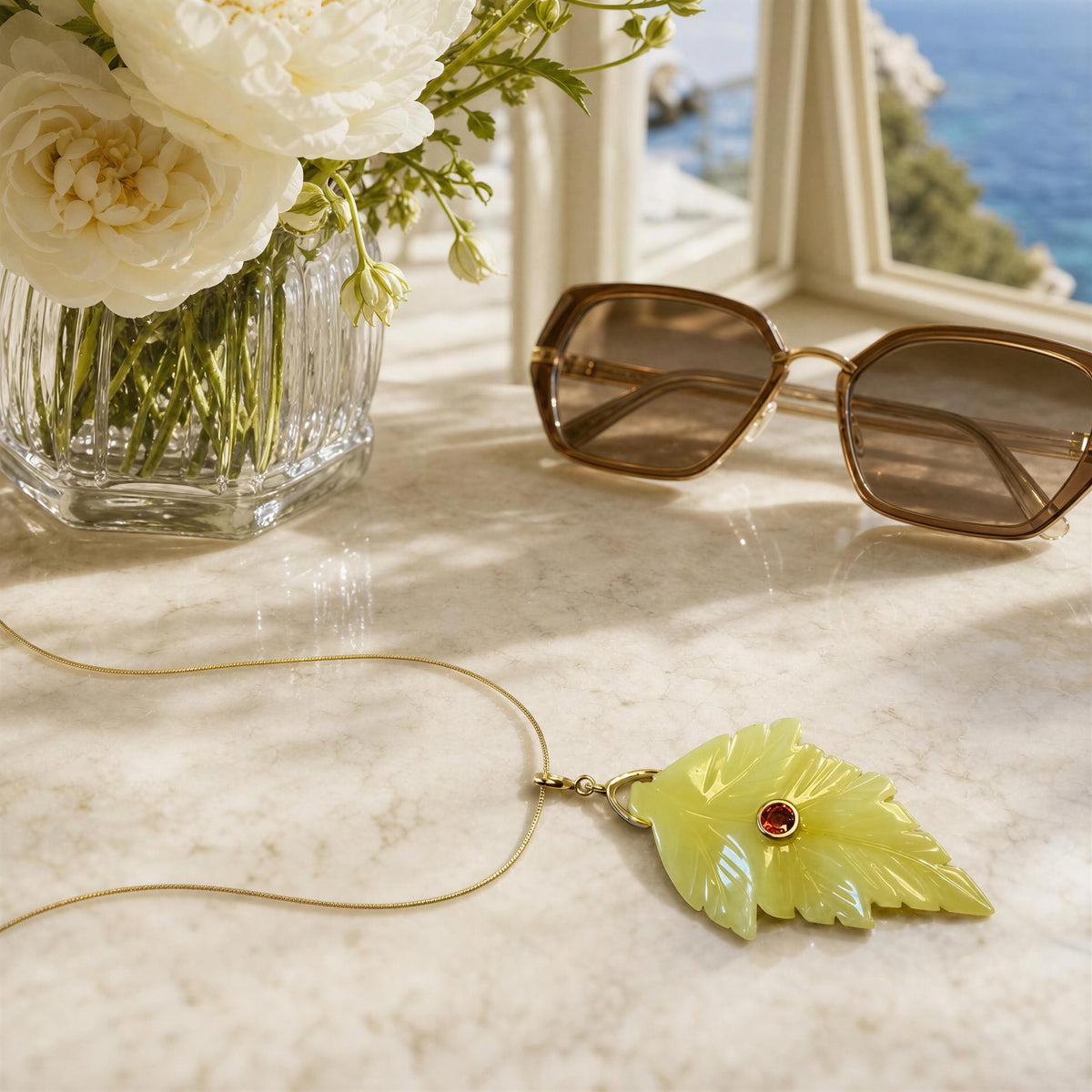 A gold necklace with a carved light green leaf pendant featuring a small red gemstone, styled on a marble surface next to sunglasses, a flower vase, and a window view.