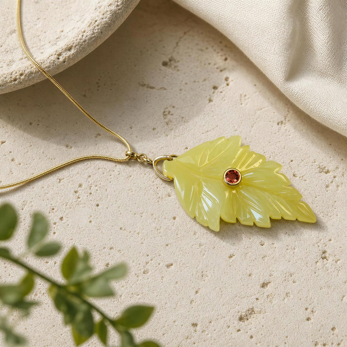 A yellow carved leaf pendant necklace with a small red gemstone and gold setting, resting on a textured beige stone surface with a cream fabric corner and blurred green leaves in the foreground.