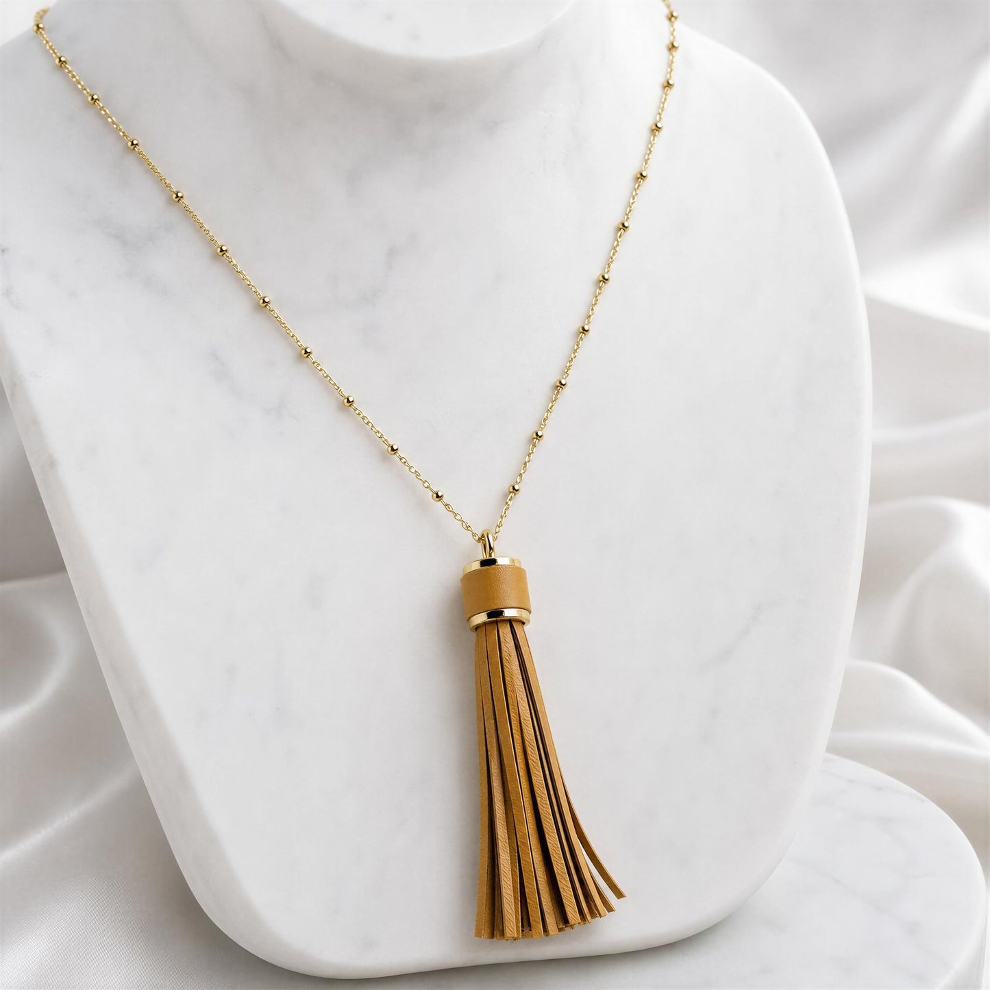 A gold chain necklace with a tan leather tassel pendant, displayed on a white marble bust.