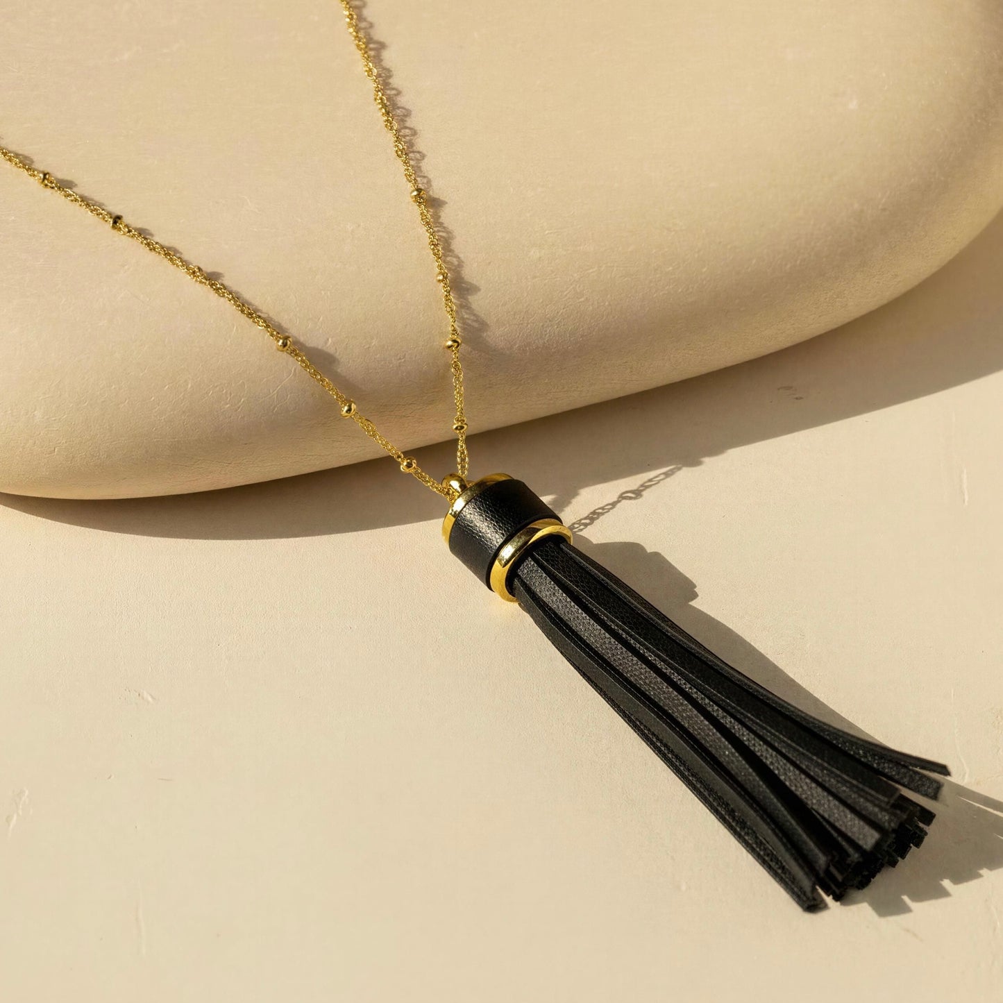 A gold chain necklace with a black leather tassel pendant, displayed on a beige surface with a matching curved prop.