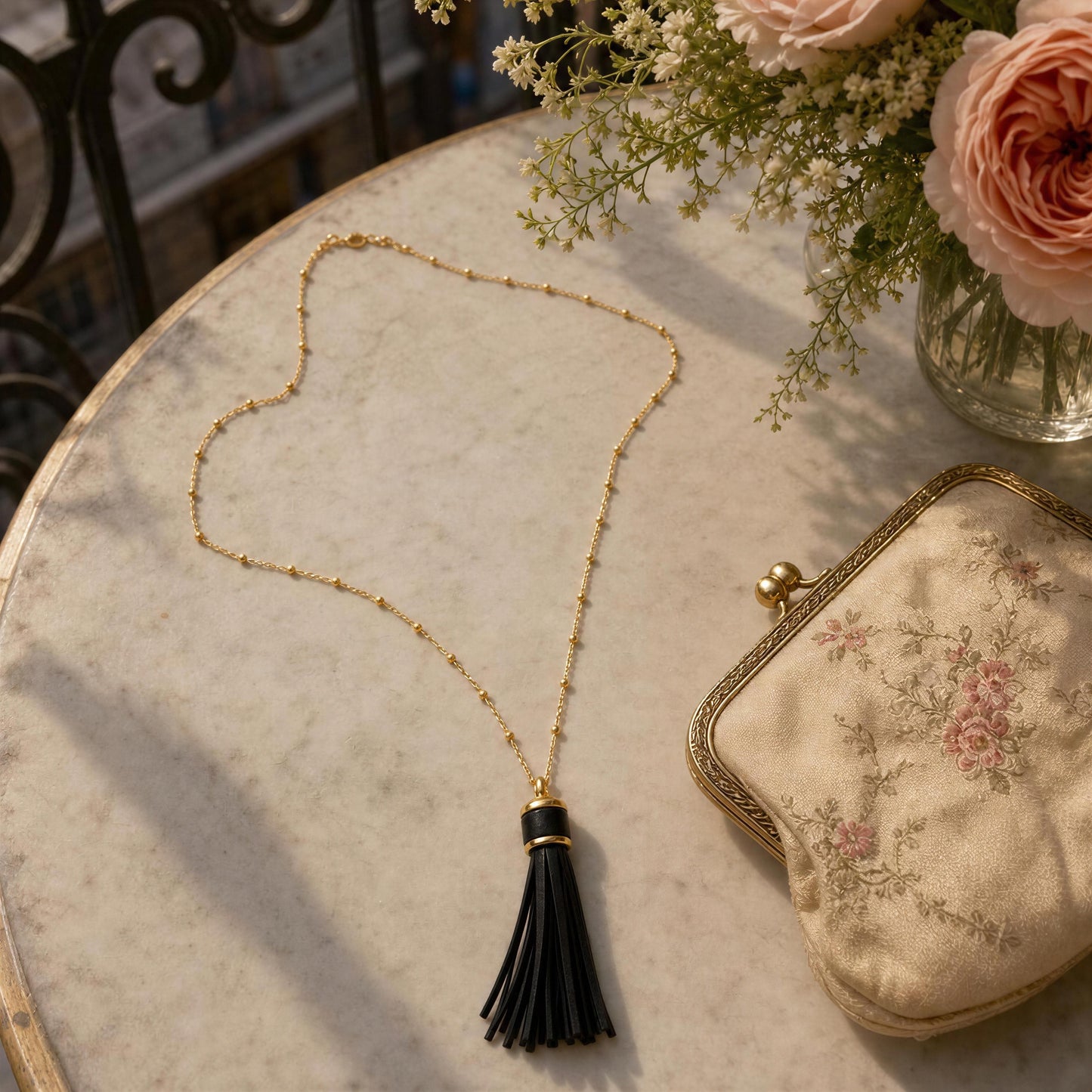 The Frangia Ebano Necklace, a gold chain necklace with a black tassel pendant is styled on a marble table alongside a floral clutch and pink roses.