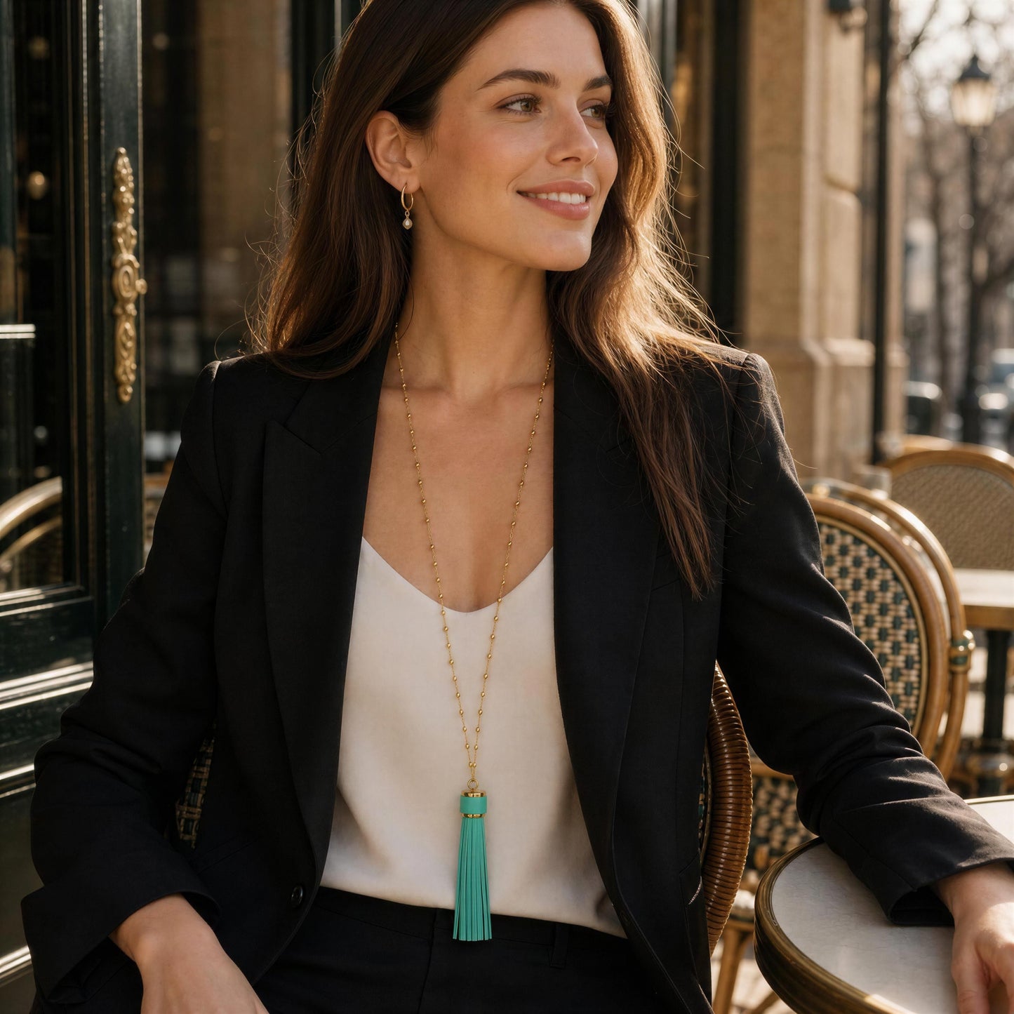 A woman wearing  the Frangia Firuze Necklace, a long turquoise tassel necklace with a delicate gold chain, styled with a black blazer and cream top, sitting at an outdoor cafe table.