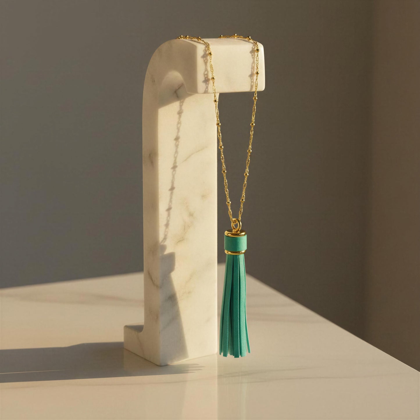 A gold chain necklace with a turquoise tassel pendant, displayed on a white marble stand against a neutral gray background.
