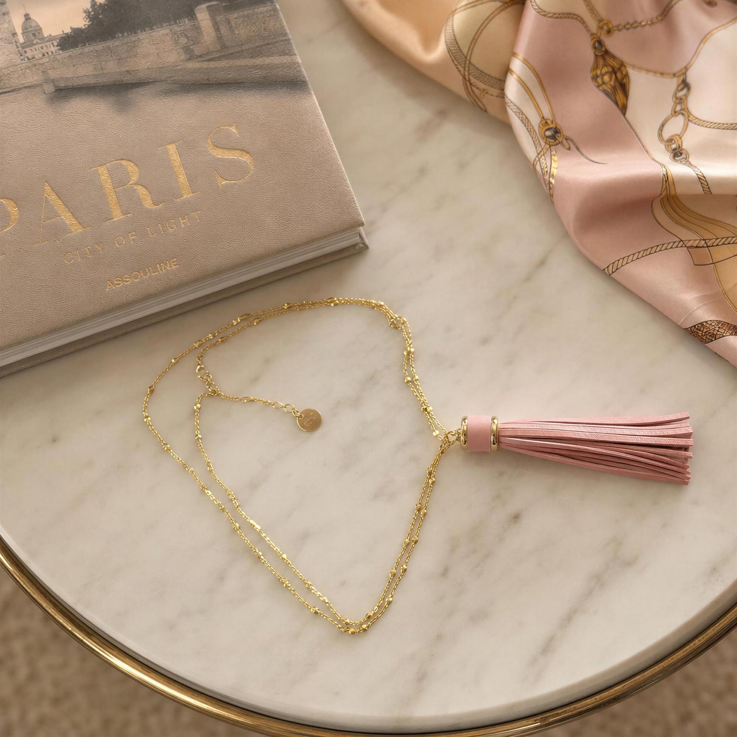 A gold chain necklace with a pink tassel pendant is arranged on a white marble tray with a gold rim. A beige book and a pink satin scarf with gold chain motifs are styled around it.