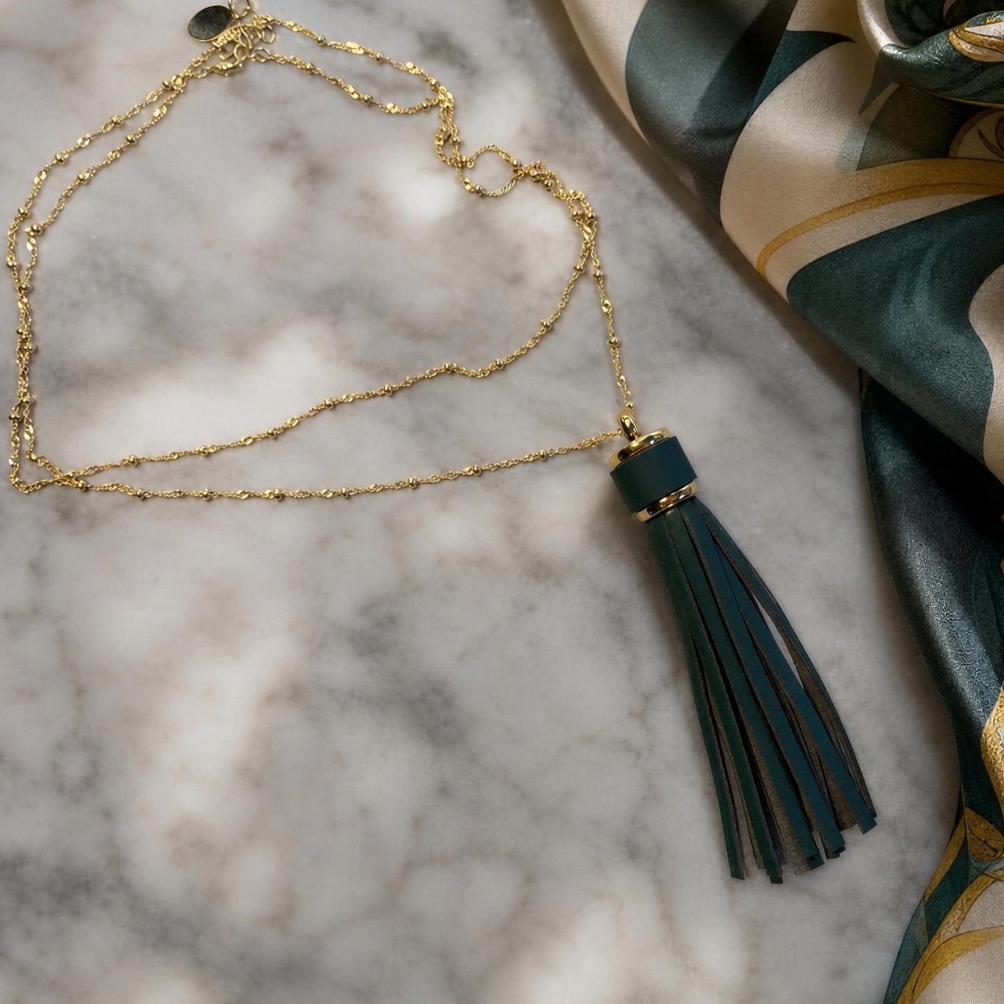 A gold chain necklace with a dark teal tassel pendant, styled on a white marble surface next to a patterned teal and gold silk scarf.