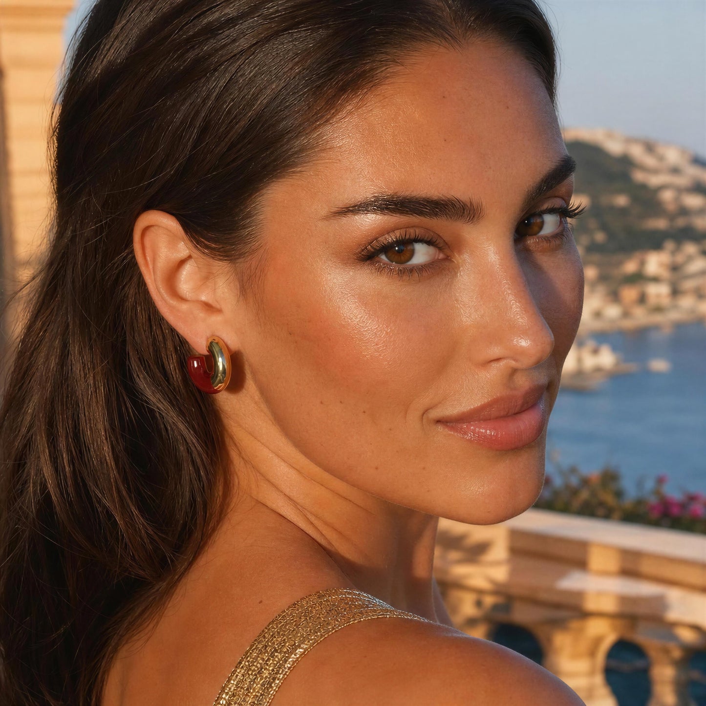 A close-up lifestyle shot of a woman with dark hair wearing the Gatsby Arc Earrings - Merlot from Chica Chica Jewelry  set against a warm coastal background.